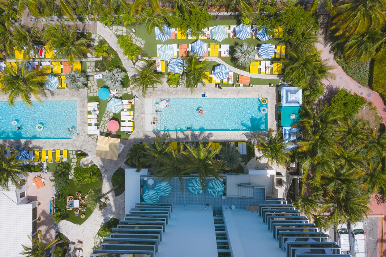 Miami Beach hotel pool deck aerial, twin pools, blue striped umbrellas, yellow loungers, palm trees