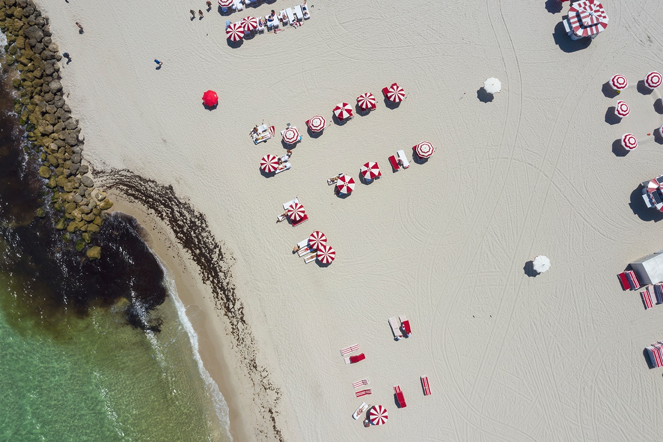 Faena Miami Beach aerial, red and white striped umbrellas on the sand, rock jetty and green Atlantic water