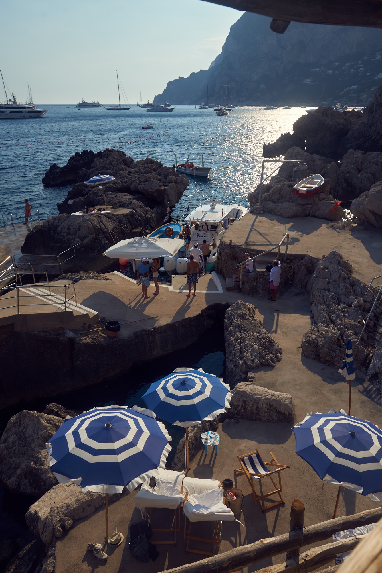 Capri beach club, blue striped umbrellas, boats moored at rocky shore, Mediterranean afternoon