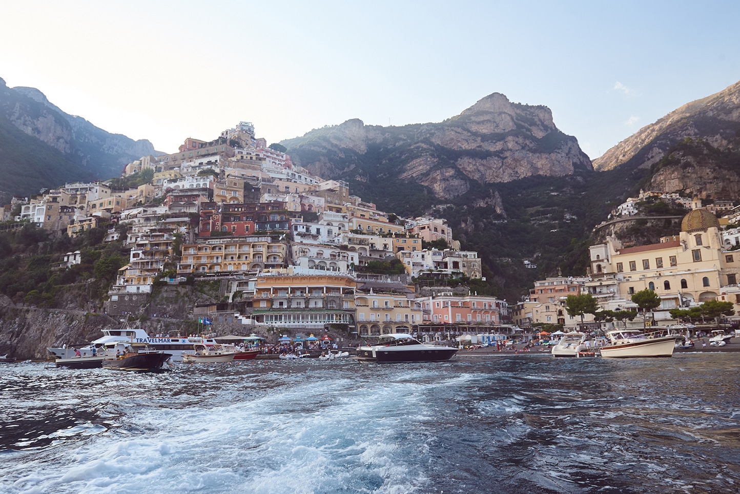 Positano from the boat, golden hour light on pastel terraced hotels, wake in foreground